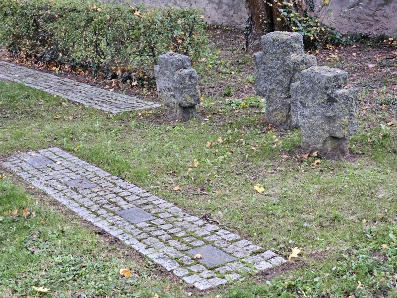 Auf dem Bild sieht man einen Rechteckigen Streifen von Pflastersteinen auf einer Rasenfläche vor drei steinernen Kreuzen. Zwischen den Pflastersteinen kann man vier größere, rechteckige, glatte Steine erkennen. Im hinteren Teil des Bildes erkennt man eine weitere, möglicherweise ähnlich Reihe von Pflastersteinen. 